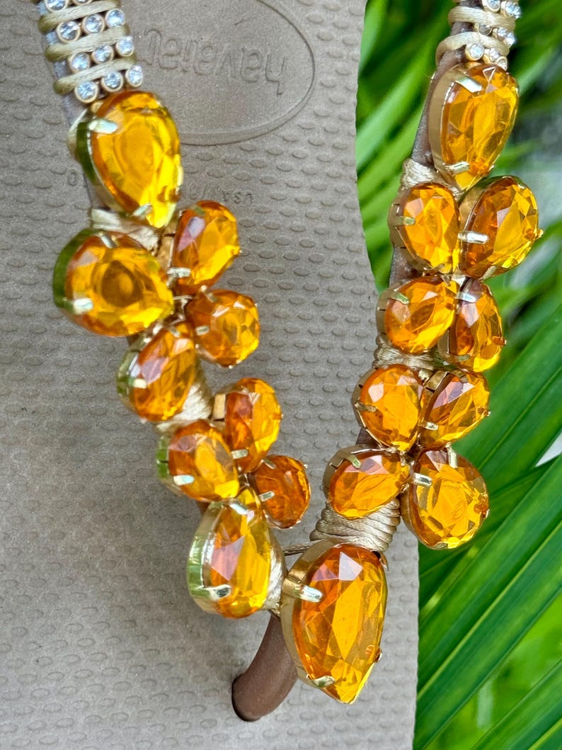 Pode incluir: Um par de chinelos marrons com uma tira dourada adornada com cristais laranja. A tira apresenta um design floral com m&uacute;ltiplos cristais.