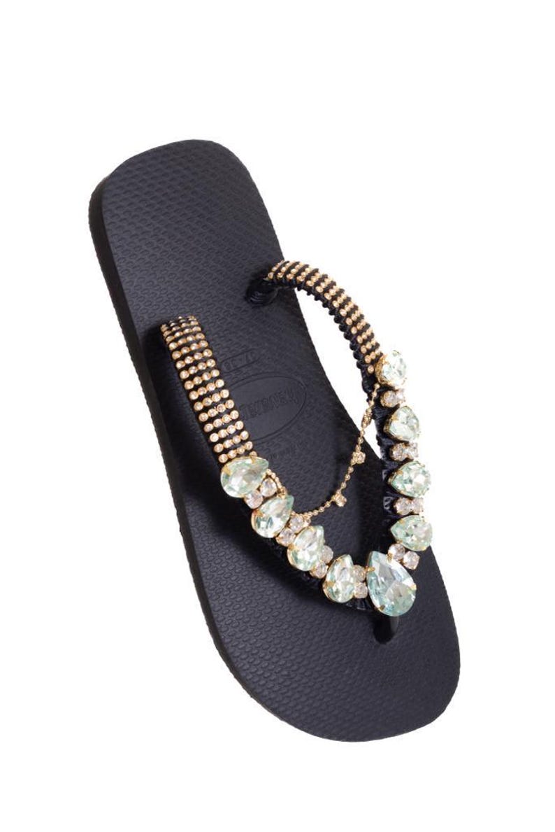 K&ouml;nnte beinhalten: Schwarze Flip-Flop-Sandale mit dekorativem Riemen. Der Riemen ist mit goldfarbenen Verzierungen und hellgr&uuml;nen und klaren Edelsteinen versehen. Der Zehenriemen ist ebenfalls mit goldfarbenen Perlen verziert. Die Sandale ist vor wei&szlig;em Hintergrund platziert.