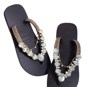 K&ouml;nnte beinhalten: Ein Paar schwarze Flip-Flops mit goldfarbenen Riemen, die mit kleinen Strasssteinen besetzt sind. Klare und hellblaue Edelsteine sind dekorativ auf den Riemen angeordnet.