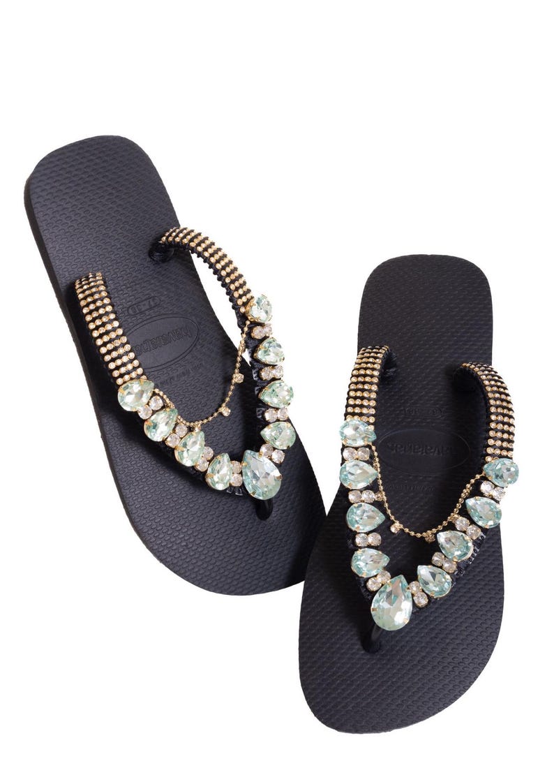K&ouml;nnte beinhalten: Ein Paar schwarze Flip-Flops mit goldfarbenen Riemen, die mit kleinen Strasssteinen besetzt sind. Klare und hellblaue Edelsteine sind dekorativ auf den Riemen angeordnet.
