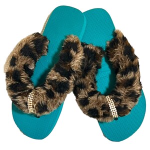 May include: A pair of teal flip flops with a brown and black leopard print fur trim and a gold rhinestone embellishment.