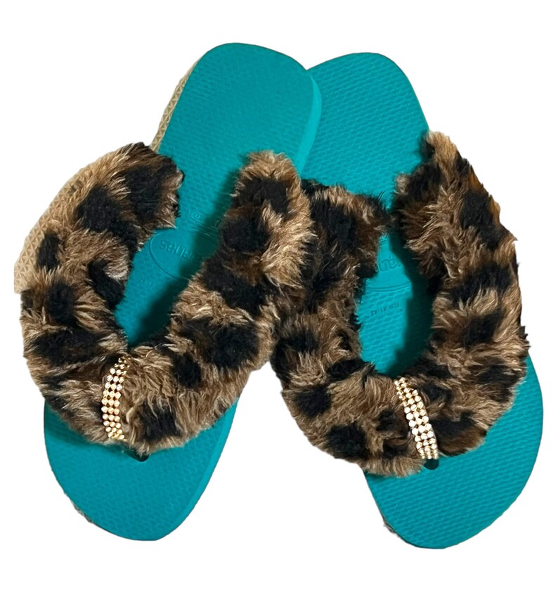 May include: A pair of teal flip flops with a brown and black leopard print fur trim and a gold rhinestone embellishment.