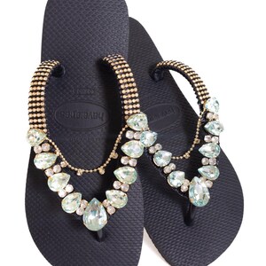 K&ouml;nnte beinhalten: Marineblaue Havaianas-Flip-Flops mit einem goldfarbenen Riemen, der mit klaren und hellblauen Edelsteinen verziert ist. Die Flip-Flops haben eine strukturierte Oberfl&auml;che und ein klassisches Zehendesign, ideal f&uuml;r den Strand.