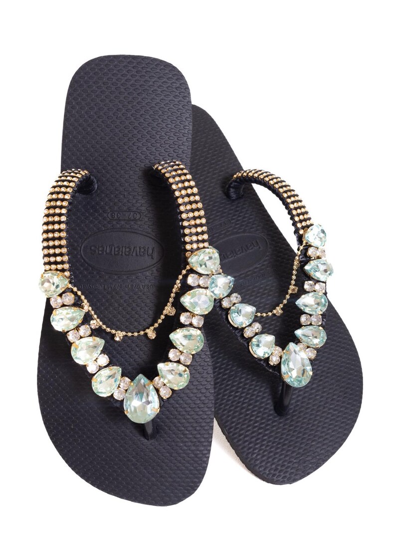 K&ouml;nnte beinhalten: Marineblaue Havaianas-Flip-Flops mit einem goldfarbenen Riemen, der mit klaren und hellblauen Edelsteinen verziert ist. Die Flip-Flops haben eine strukturierte Oberfl&auml;che und ein klassisches Zehendesign, ideal f&uuml;r den Strand.