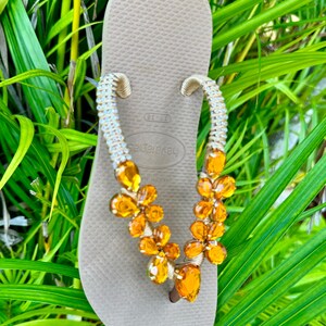 Pode incluir: Chinelos bege com uma tira de contas adornada com strass laranja em forma de flor.