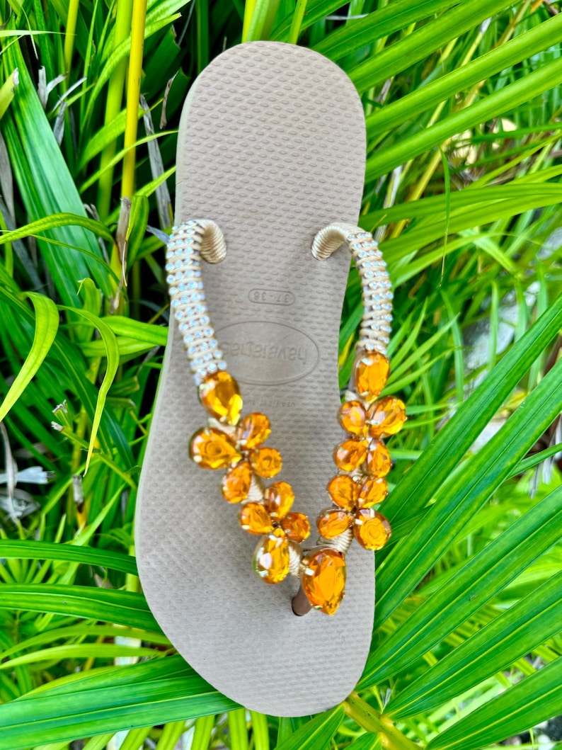 Pode incluir: Chinelos bege com uma tira de contas adornada com strass laranja em forma de flor.