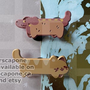 May include: Two enamel pins featuring stylised animals. The top pin is a brown dog with a hot dog bun body. The bottom pin is a tan cat with a long body. Text on the image reads: marscapone only available on marscapone.ca and etsy.