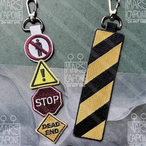 May include: Two embroidered keychains with metal lobster clasps. One features a vertical strip with black and yellow diagonal hazard stripes. The other consists of four stacked embroidered patches: a no-pedestrian sign, a yellow warning triangle, a red stop sign, and a yellow diamond sign with the text DEAD END.