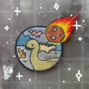 May include: Embroidered patch featuring a cartoon dinosaur scene with a meteor. The patch has a blue border, a yellow dinosaur, and a brown meteor with red and yellow flames. The background is a starry sky.