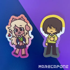 Puede incluir: Dos pegatinas troqueladas con personajes de dibujos animados. La pegatina de la izquierda muestra un personaje de pelaje claro con astas, un suéter verde y rojo y una falda oscura sobre una bandera lésbica. La de la derecha muestra un personaje de pelo castaño con un suéter amarillo sobre una bandera no binaria. El texto "MARSCAPONE" está en la parte inferior.