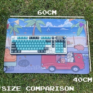 May include: A rectangular mousepad with a beach scene design, measuring 60cm long and 40cm wide. A teal and white keyboard sits on the pad. The pad features a red car with money bags and the text "CAPITALISM".