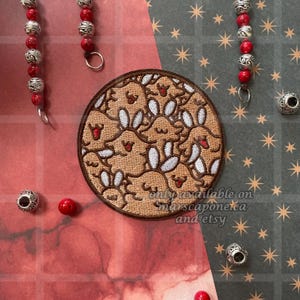 May include: A circular embroidered patch featuring a repeating pattern of cartoon bunnies in shades of brown and white. The patch has a brown border and is set against a red and green background with decorative beads.