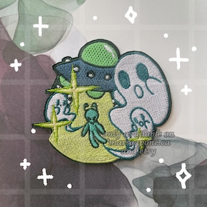 Może przedstawiać: Haftowana naszywka z fantazyjnym wzorem zielono-niebieskiego stworzenia przypominającego kosmitę, dwóch duchów i gwiazd. Naszywka ma jasną zieleń, niebieski i biały schemat kolorów. Tekst na naszywce brzmi "only available on marsiapone.ca and etsy."