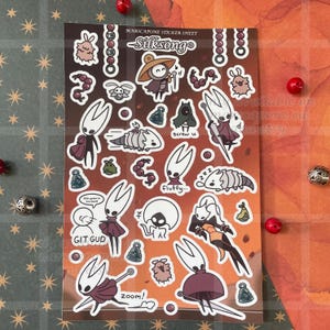 May include: A rectangular sticker sheet titled MARSCAPONE STICKER SHEET Silksong featuring various cartoon character illustrations, including white horned figures in red cloaks, small fluffy creatures, bells, and text reading GIT GUD and screw u.
