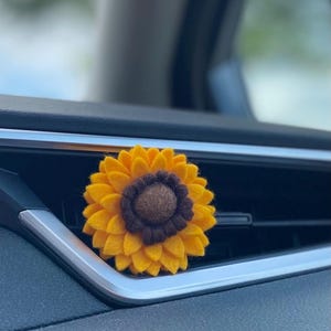 May include: A felt sunflower car air vent clip. The flower features bright yellow petals and a dark brown centre. The clip is attached to a car vent, adding a decorative touch to the vehicle's interior.