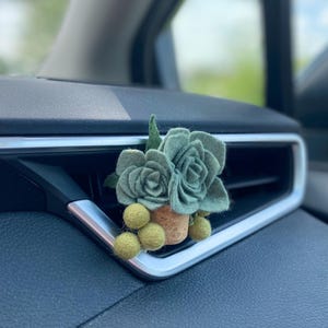May include: A sage green felt succulent air vent clip for a car. The handmade decoration features felt flowers and small yellow-green felt balls, set in a small cork pot. The clip is designed to attach to a car's air vent.
