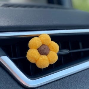 May include: A yellow felt flower car air vent clip with a brown centre. The flower is made of soft, round petals and is designed to clip onto a car's air vent. The car's dashboard is black.