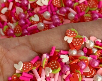 May include: A mix of pink, red, white, and green sprinkles in the shape of hearts, strawberries, and small cylinders. The sprinkles are in a hand.
