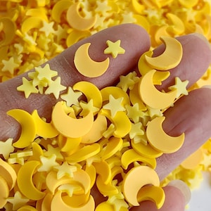 May include: A close-up of a pile of yellow and orange resin moon and star shapes. The shapes are scattered on a hand.
