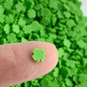 May include: A close-up of a green four-leaf clover sprinkle. The sprinkle is small and is being held between a finger and thumb. The background is a pile of similar green four-leaf clover sprinkles.