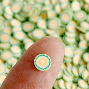 May include: A close-up of a small, yellow and green circle with a white center. The circle is surrounded by a white border with a green and yellow pattern. The circle is being held by a finger.