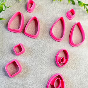 May include: A collection of bright pink polymer clay cutters in various shapes, including teardrops, ovals, and squares. The cutters are arranged on a neutral-toned surface, with green foliage in the background. These tools are likely for crafting or baking.