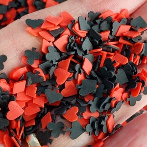 May include: A close-up of red and black card suit sprinkles. The sprinkles are in the shape of hearts, diamonds, clubs, and spades.