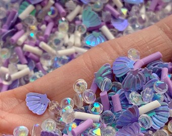 May include: A mix of purple, white, and iridescent sprinkles, including seashells, beads, and small cylinders.