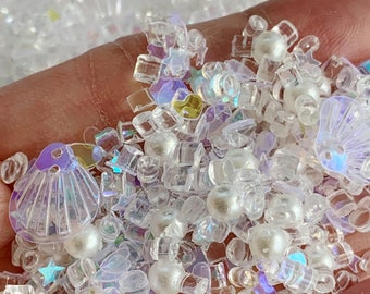 May include: A mix of iridescent clear plastic and white pearl beads, seashells, and stars. The beads and shells are in various shapes and sizes.