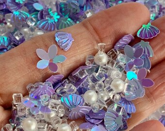 May include: A handful of iridescent craft supplies including seashell and flower-shaped sequins, clear cube beads, and white pearl beads. The color palette includes shades of purple, blue, and white, creating a shimmering effect.