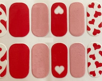 May include: Nail wraps featuring a variety of designs. Some wraps are solid red or pink, while others have a white background with red hearts. One red wrap has a white heart cutout.
