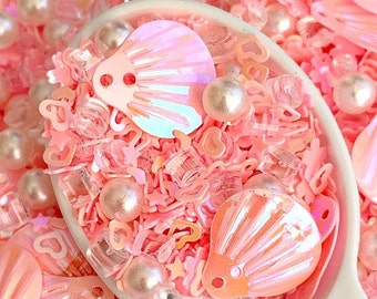 May include: A white spoon filled with pink and iridescent glitter, seashells, and pearls. The glitter is in various shapes, including stars, hearts, and circles.