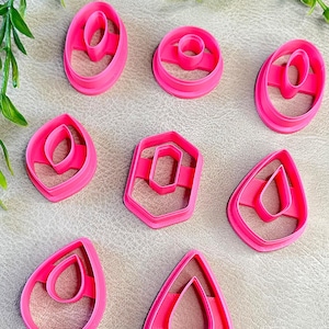 May include: A collection of bright pink polymer clay earring cutters in various shapes, including ovals, teardrops, and a hexagon. The cutters are arranged on a neutral surface, with green foliage in the background. These tools are for crafting earrings.