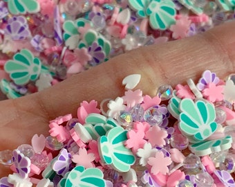 May include: A mix of colorful resin sprinkles for crafting. The sprinkles are shaped like seashells, flowers, and hearts. There are also clear iridescent beads.