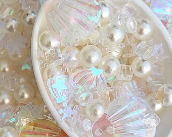 May include: A white scoop filled with iridescent seashell and pearl-like beads. The beads are a mix of white and iridescent colors. The seashells are iridescent and have a holographic finish.