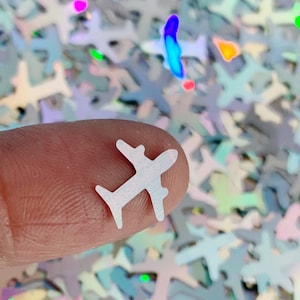 May include: White iridescent glitter shaped like airplanes. The glitter is scattered on a surface and a finger is holding one of the glitter pieces.