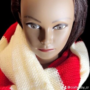 May include: A knitted scarf in a repeating pattern of red and cream. The scarf is wrapped around a mannequin head with brown hair and a neutral skin tone. The background is black.