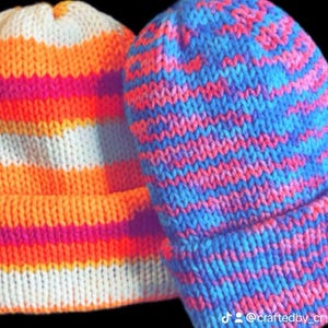 May include: Two double-sided beanies. One beanie features horizontal stripes of white, orange, and pink. The other beanie is blue with pink stripes. The beanies are handmade and knitted.