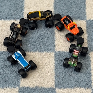May include: A collection of five die-cast monster trucks arranged in a semi-circle. The vehicles include a gold Earth Shaker, an orange truck, a black and silver Grave Digger with green flames, a blue and silver truck, and a black patterned truck.