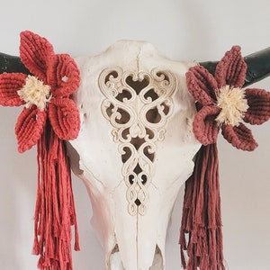 May include: White cow skull with intricate carvings, decorated with two red crocheted flowers with tassels.