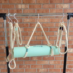 May include: A light green yoga mat with the word "Gaiam" printed on it is hanging from a white metal rack with a white rope strap. The mat is hanging with other white rope straps.