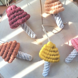 May include: Five handmade crocheted mushroom ornaments in various colors, including red, orange, and yellow. The mushrooms are made with a thick, textured yarn and have a natural rope stem.
