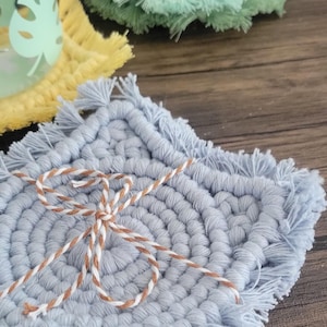 May include: A collection of handmade macrame coasters in various colors, including blue, yellow, and green. The coasters have a fringed edge and are tied together with twine. The coasters are on a wooden surface.