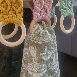 May include: Three crocheted towel rings in yellow, pink, and green hanging on a silver stove. A green towel is hanging from the pink ring.