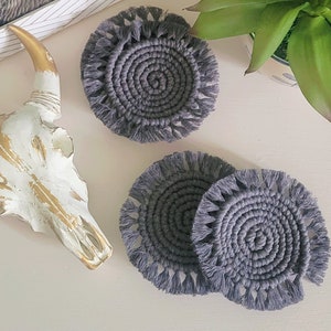 May include: Three gray crocheted coasters with fringe. The coasters are round and have a circular pattern in the center.