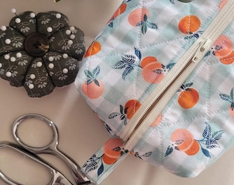 May include: A floral pin cushion, a peach-patterned quilted pouch with a zipper, and silver scissors are arranged on a white surface. The pouch features a light blue and white gingham pattern with orange peaches and green leaves. The pin cushion is dark green with white dots.