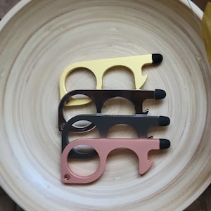 May include: Four door openers in a wooden bowl. The door openers are in gold, black, rose gold, and silver. Each opener has a loop, a stylus, and a bottle opener.