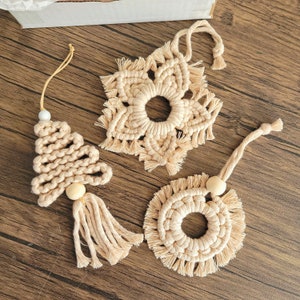 May include: Three handmade macrame ornaments in the shape of a snowflake, a Christmas tree, and a circle. Each ornament is made with natural beige cotton cord and features a wooden bead.