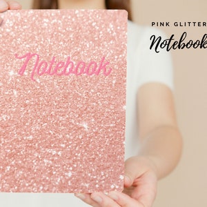 May include: A pink glitter notebook with the word "Notebook" in pink script. The notebook is held by a person. The background is a light beige colour. The notebook is a rectangular shape.