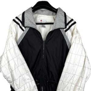 May include: A vintage Avon jacket in black, white, and houndstooth patterns. The jacket features a full zipper, a drawstring waist, and a collared neckline. The sleeves are white with a grid pattern, and the shoulders have black stripes.